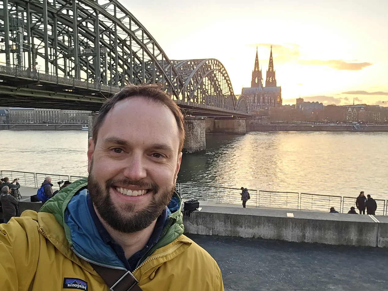 Justin Fuhrmann on the Rhine riverfront in Cologne, the Hohenzollern Bridge and Cologne Cathedral visible behind him at sunset